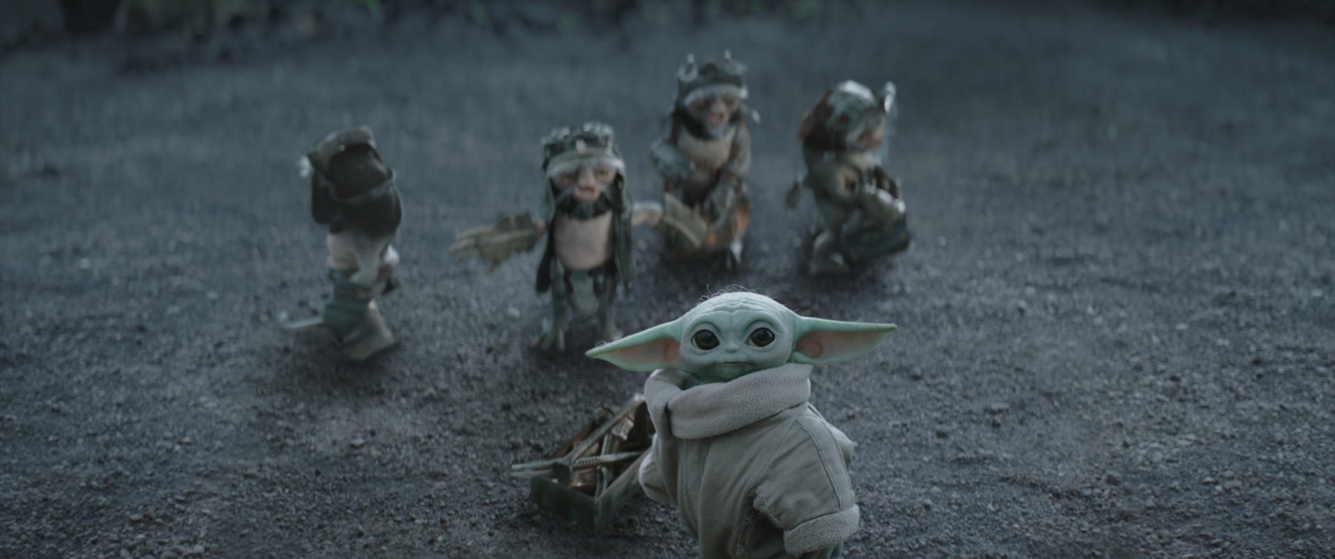Baby-Yoda in einem grauen Mantel blickt nach vorne, während vier kleine Kreaturen im Hintergrund auf sandigem Boden stehen.