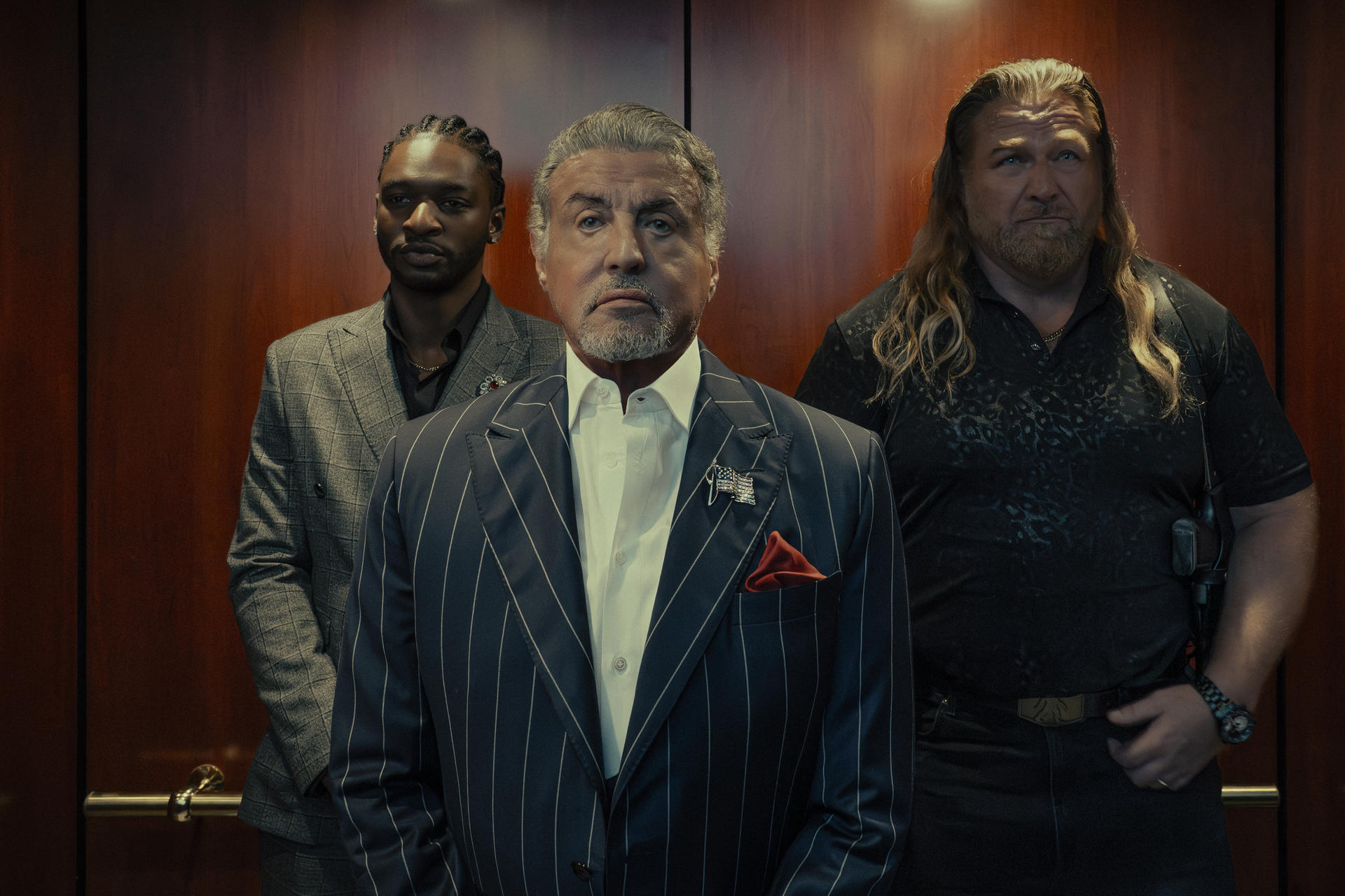 Drei Männer stehen in einem Aufzug mit holzverkleideten Wänden; Sylvester Stallone steht im Nadelstreifenanzug in der Mitte.