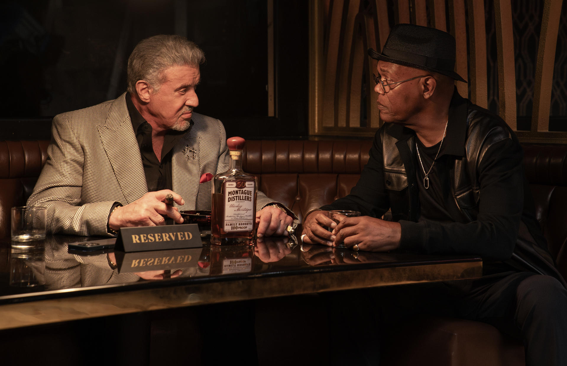 Sylvester Stallone und Samuel L. Jackson sitzen in einer Bar an einem Tisch mit einer Flasche Montague Distillers Whiskey.