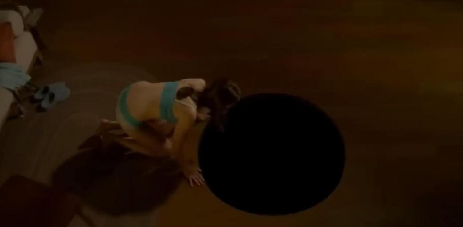 Eine Frau kniet auf einem dunklen Holzboden neben einem runden schwarzen Loch.