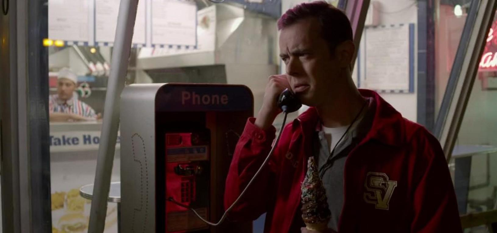 Ein Mann in roter Jacke hält nachts vor einem Laden ein Eis und einen Telefonhörer.