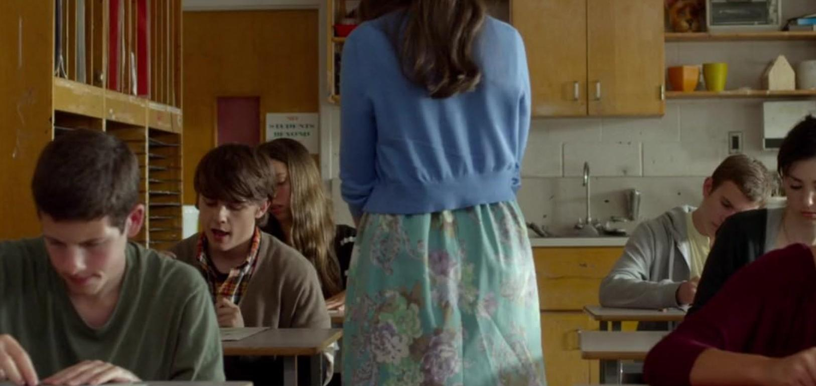 Eine Frau in blauem Oberteil und gemustertem Rock steht vor Schülern in einem Klassenzimmer.