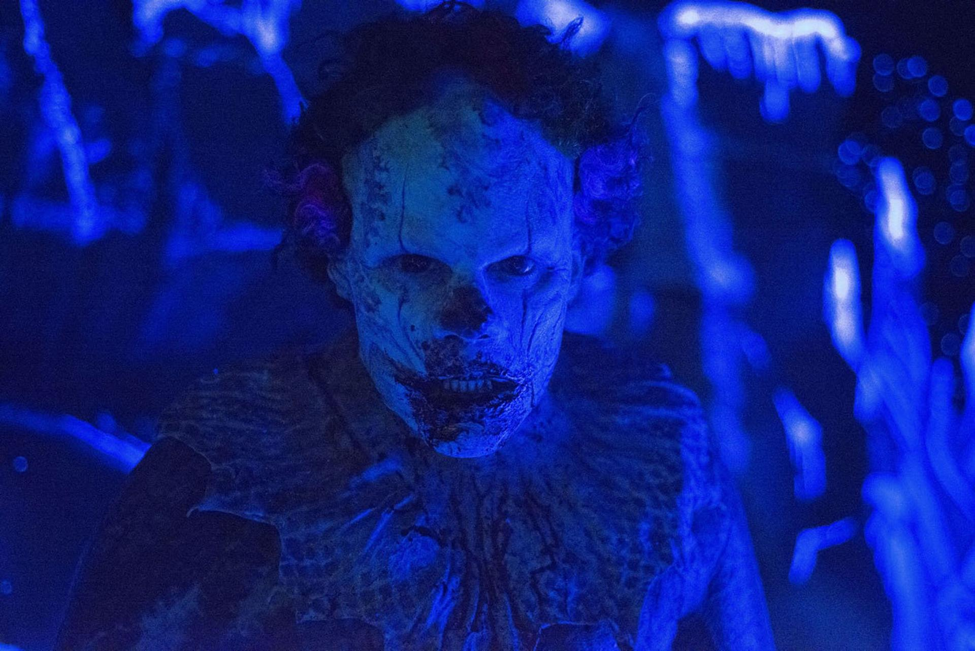 Ein gruseliger Clown mit blutverschmiertem Gesicht blickt unter blauem Licht direkt in die Kamera.