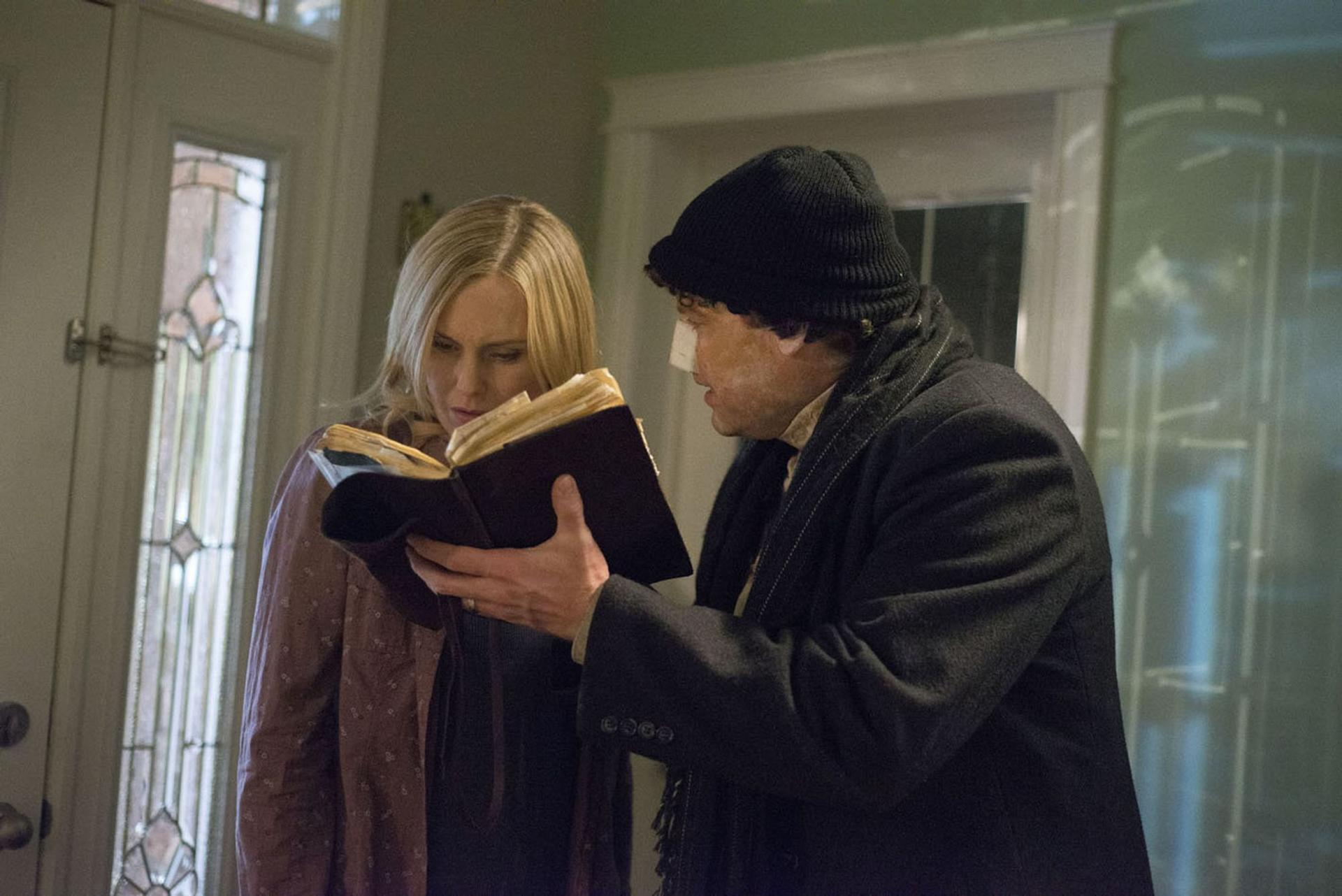 Eine blonde Frau und ein Mann mit Mütze schauen zusammen in ein altes Buch in einem Innenraum.