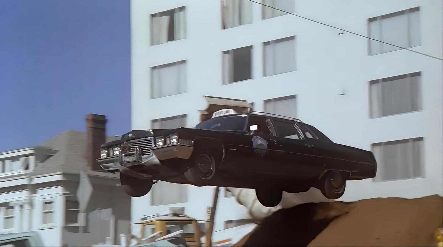 Ein schwarzes Oldtimer-Auto fliegt durch die Luft über einen Schmutzhügel vor einem weißen Gebäude.