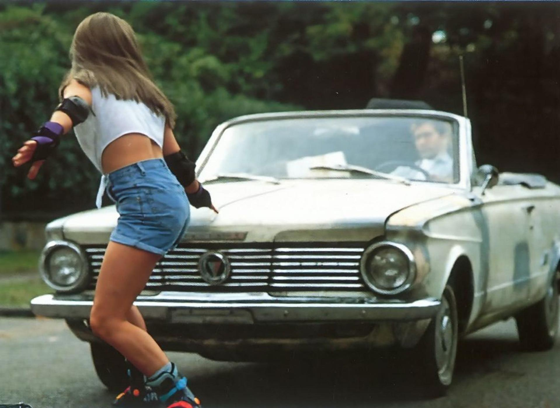 Ein Mädchen mit Inlineskates und Schutzkleidung beugt sich vor ein parkendes Cabrio.