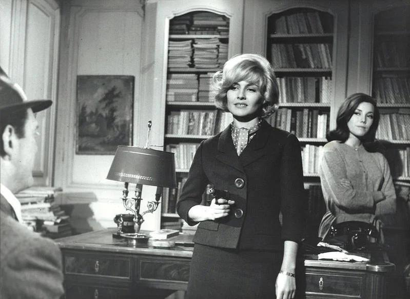 Eine blonde Frau im dunklen Kostüm hält in einem Arbeitszimmer mit Bücherregalen eine Pistole.
