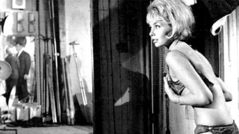 Eine blondhaarige Frau in Unterwäsche schaut in einem dunklen Raum mit Filmrequisiten im Hintergrund zur Seite.