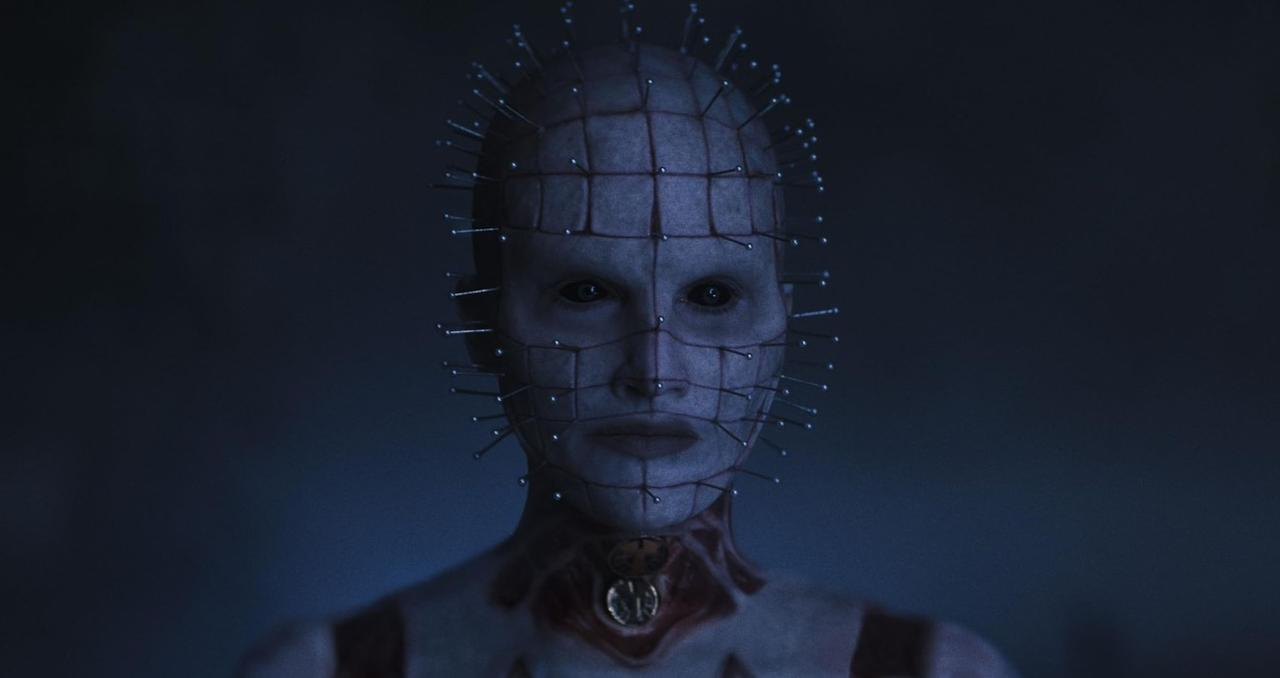 Close-up of a pale figure with pins piercing its head and face in a dark setting.