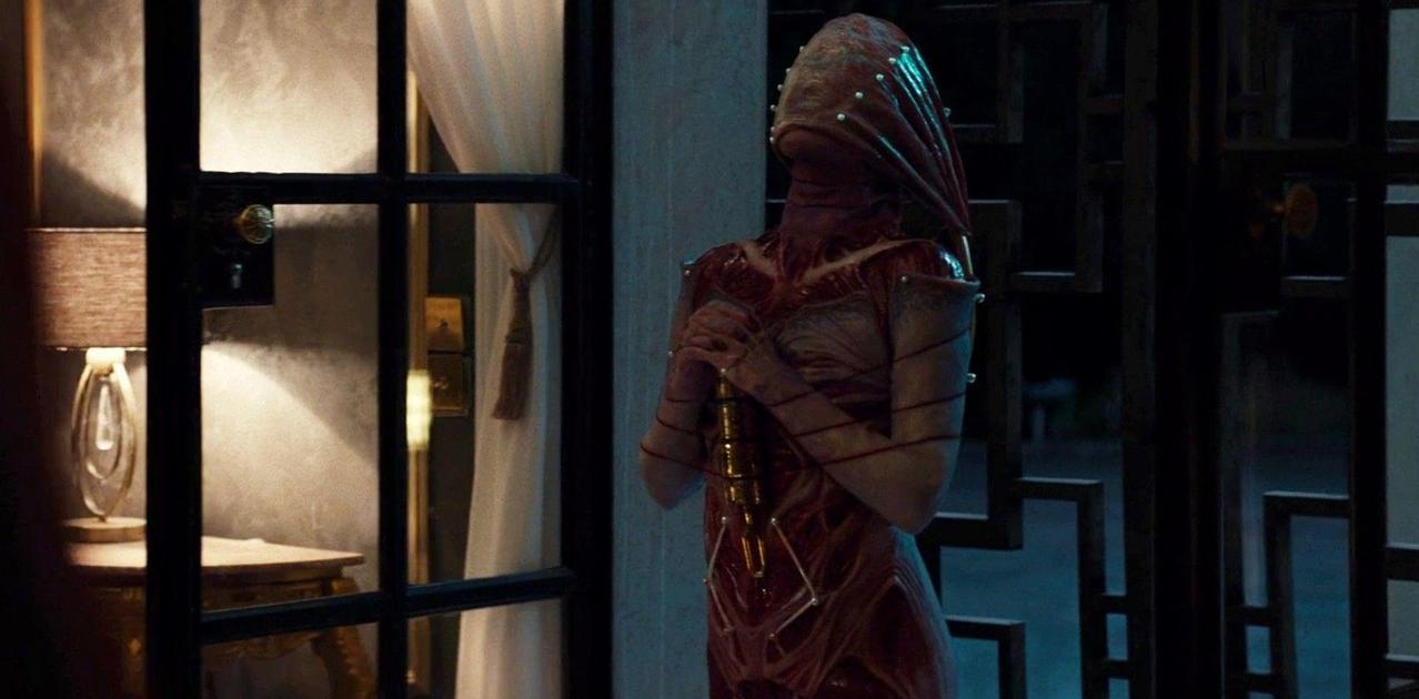 Eine humanoide Figur in einem rötlichen Anzug mit Kapuze hält drinnen ein Metallobjekt.