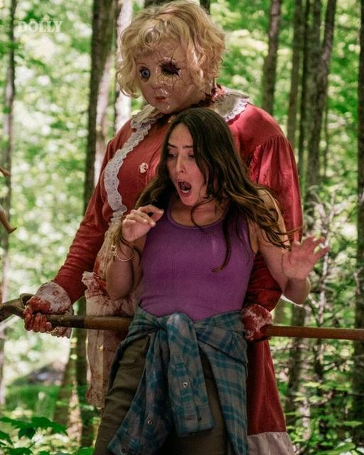 Eine erschrockene Frau in einem Wald mit einer maskierten Figur im Hintergrund, die ein blutiges Werkzeug hält.