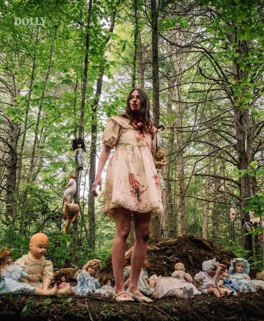 Eine blutige Frau in einem Kleid steht im Wald, umgeben von alten Puppen.