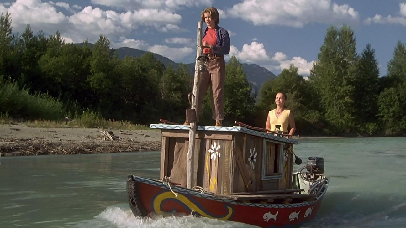 Zwei Frauen fahren auf einem bunt bemalten Holzhoot auf einem Fluss durch eine waldreiche Landschaft.