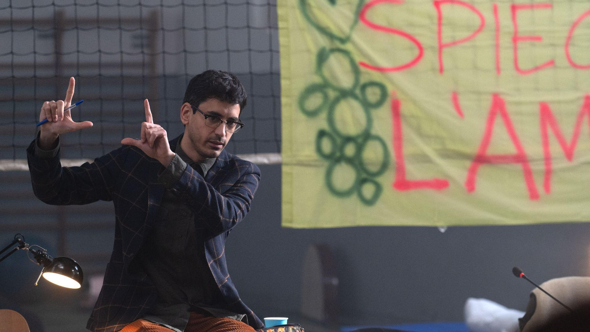 Ein Mann mit Brille und Karo-Blazer gestikuliert vor einem gelben Banner mit roter und grüner Aufschrift.