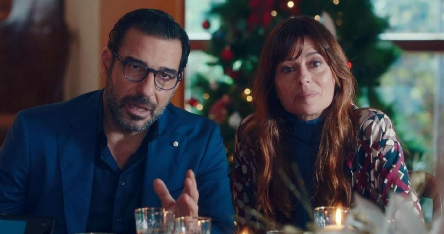 Ein Mann mit Brille und Anzug spricht, während eine Frau mit langem Haar neben ihm sitzt; im Hintergrund ein Weihnachtsbaum.
