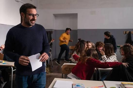 Ein lächelnder Lehrer hält in einem Klassenzimmer ein Papier, während Schüler an Tischen sitzen und ein Junge vorbeiläuft.