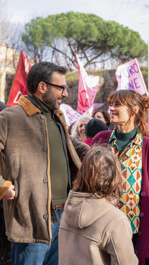 Ein Mann und eine Frau lächeln sich im Freien an, mit einem Kind im Vordergrund und rosa Protestbannern dahinter.