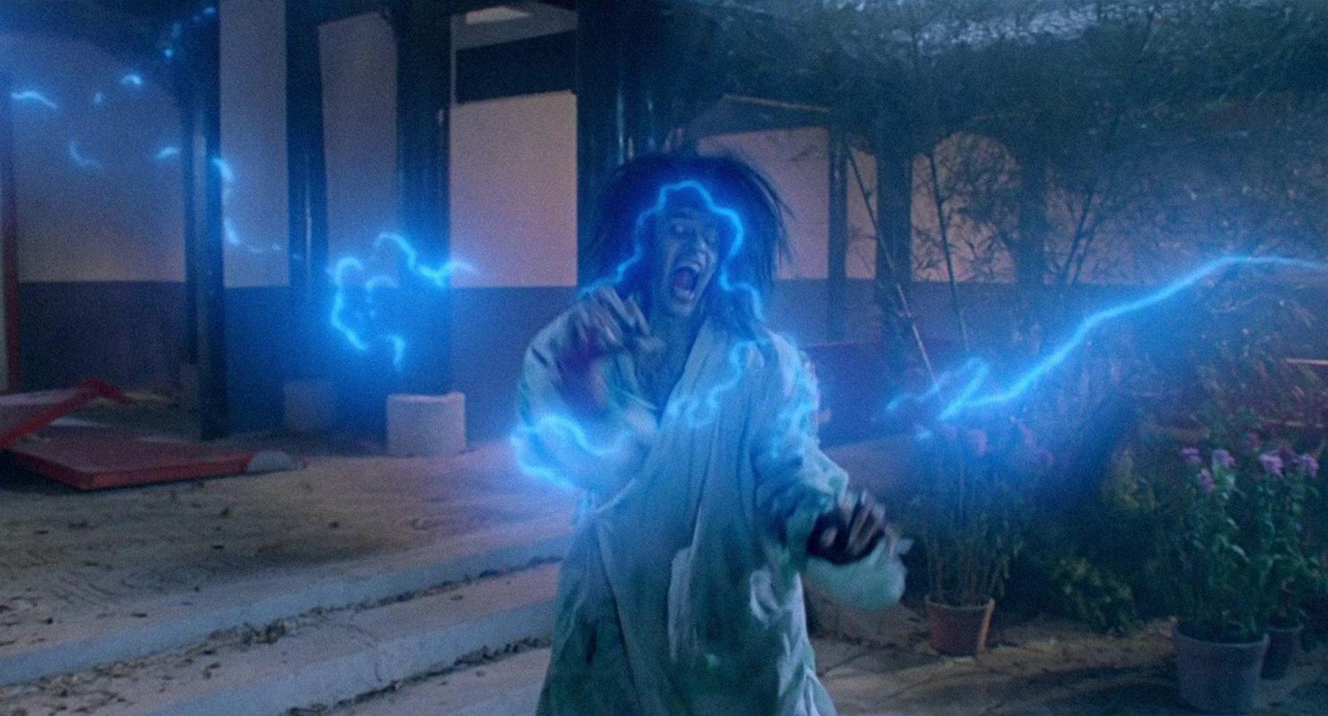 Eine Person im weißen Gewand schreit, umgeben von blauen elektrischen Blitzen vor einem Gebäude und Pflanzen.