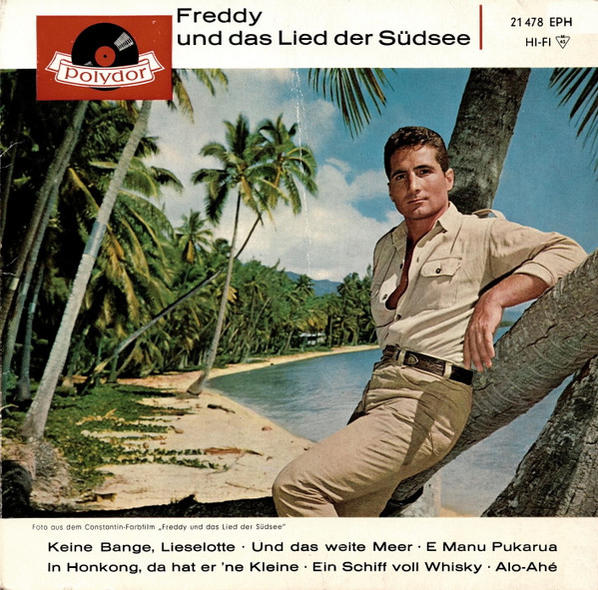 Ein Mann in Khakikleidung lehnt an einer Palme an einem tropischen Strand mit blauem Meer.