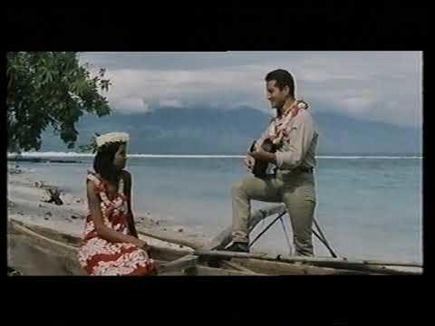Ein Mann spielt Ukulele für eine Frau am Strand mit Meer und Bergen im Hintergrund.