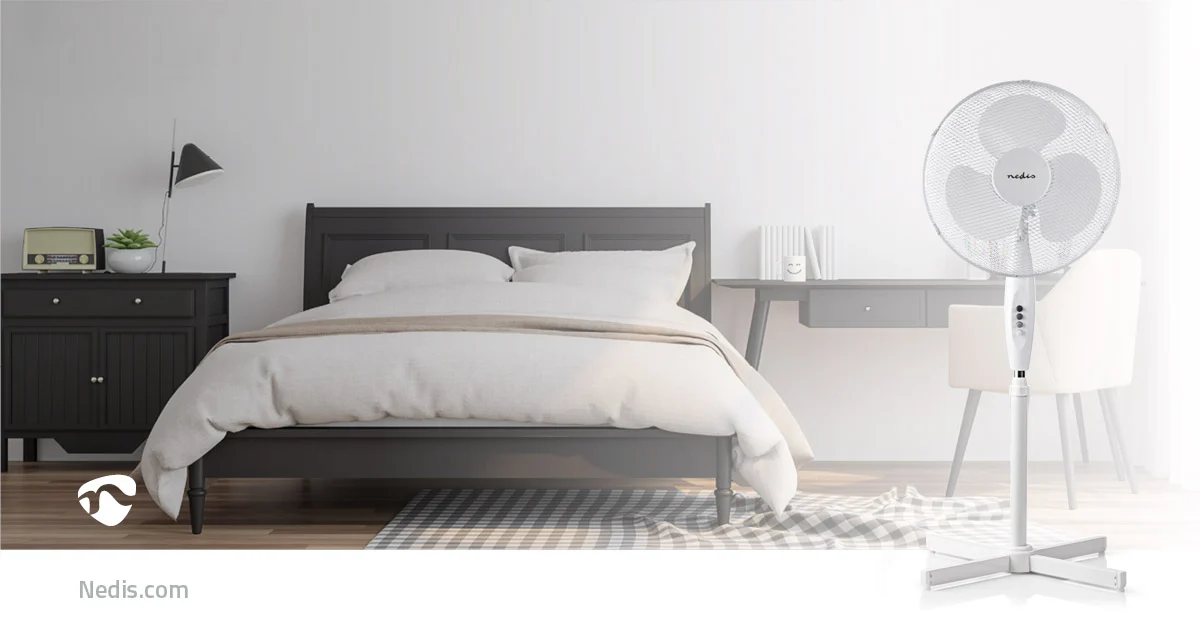 Ein weißer Standventilator steht vor einem gut eingerichteten Schlafzimmer mit einem dunklen Bett.