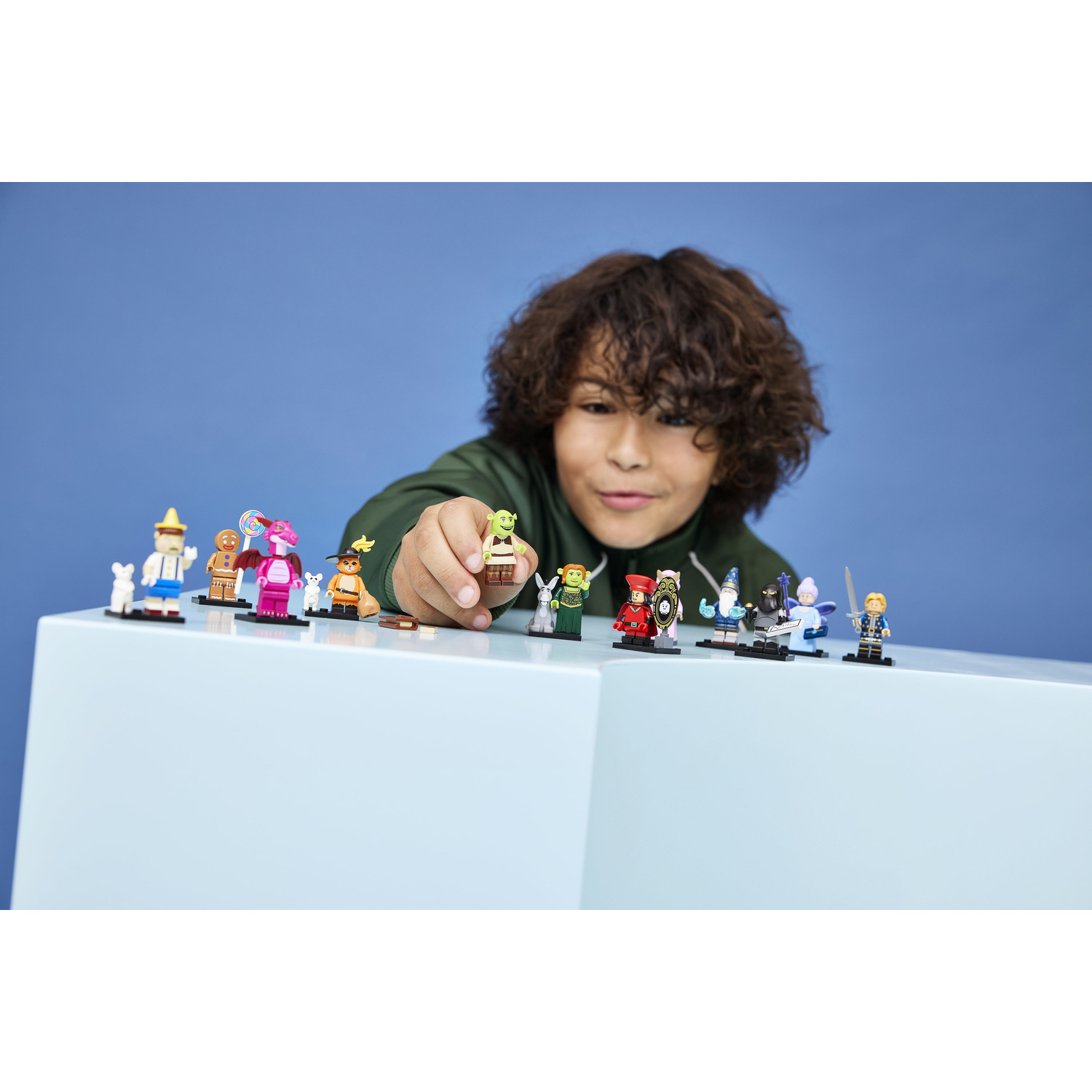 Un bambino tiene in mano una piccola minifigura Lego di Shrek davanti a un muro blu, circondato da altre figure.