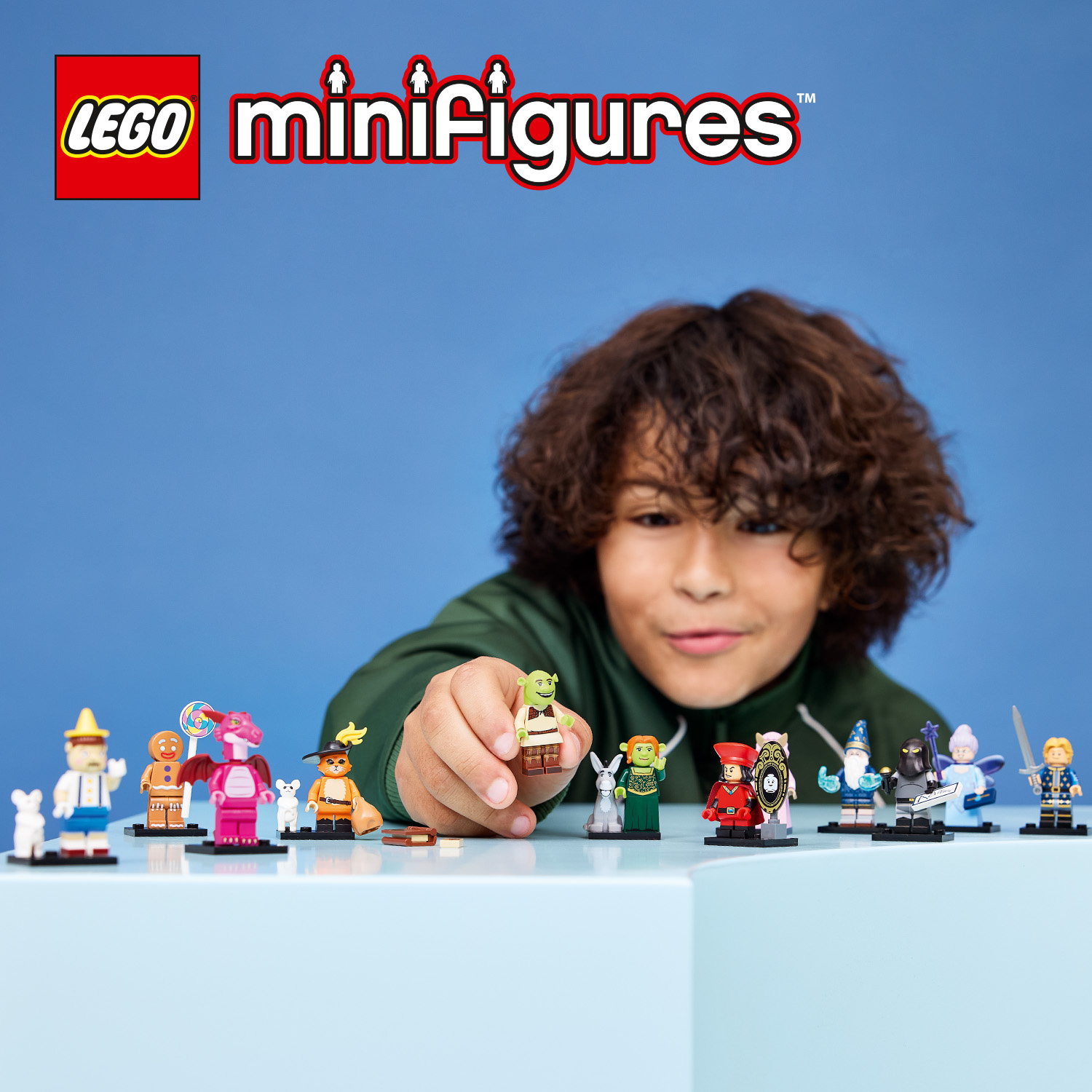 Un bambino tiene in mano una minifigura LEGO di Shrek davanti a un muro blu con altre minifigure di Shrek sul tavolo.