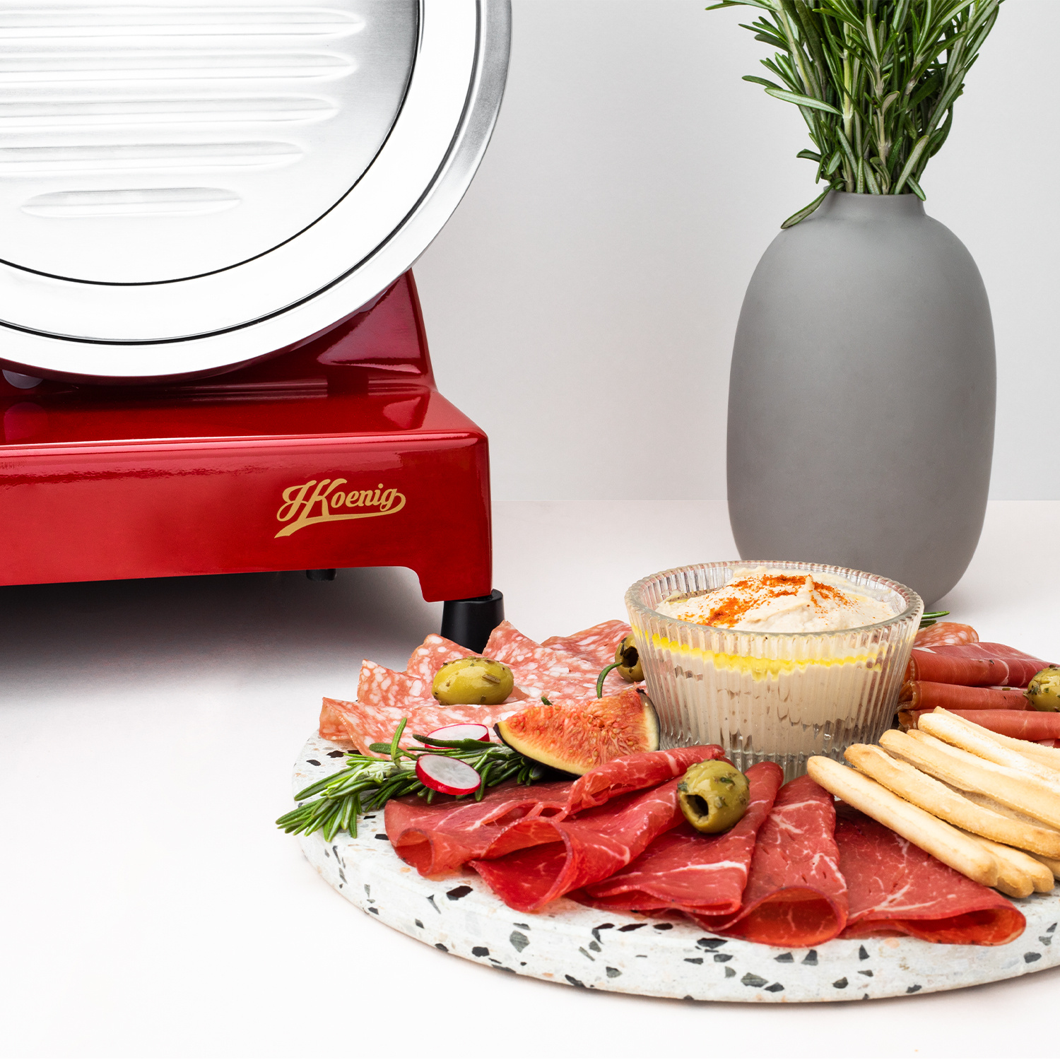 Un'affettatrice rossa si trova accanto a un piatto di marmo con salumi, salsa, grissini, olive e un vaso.