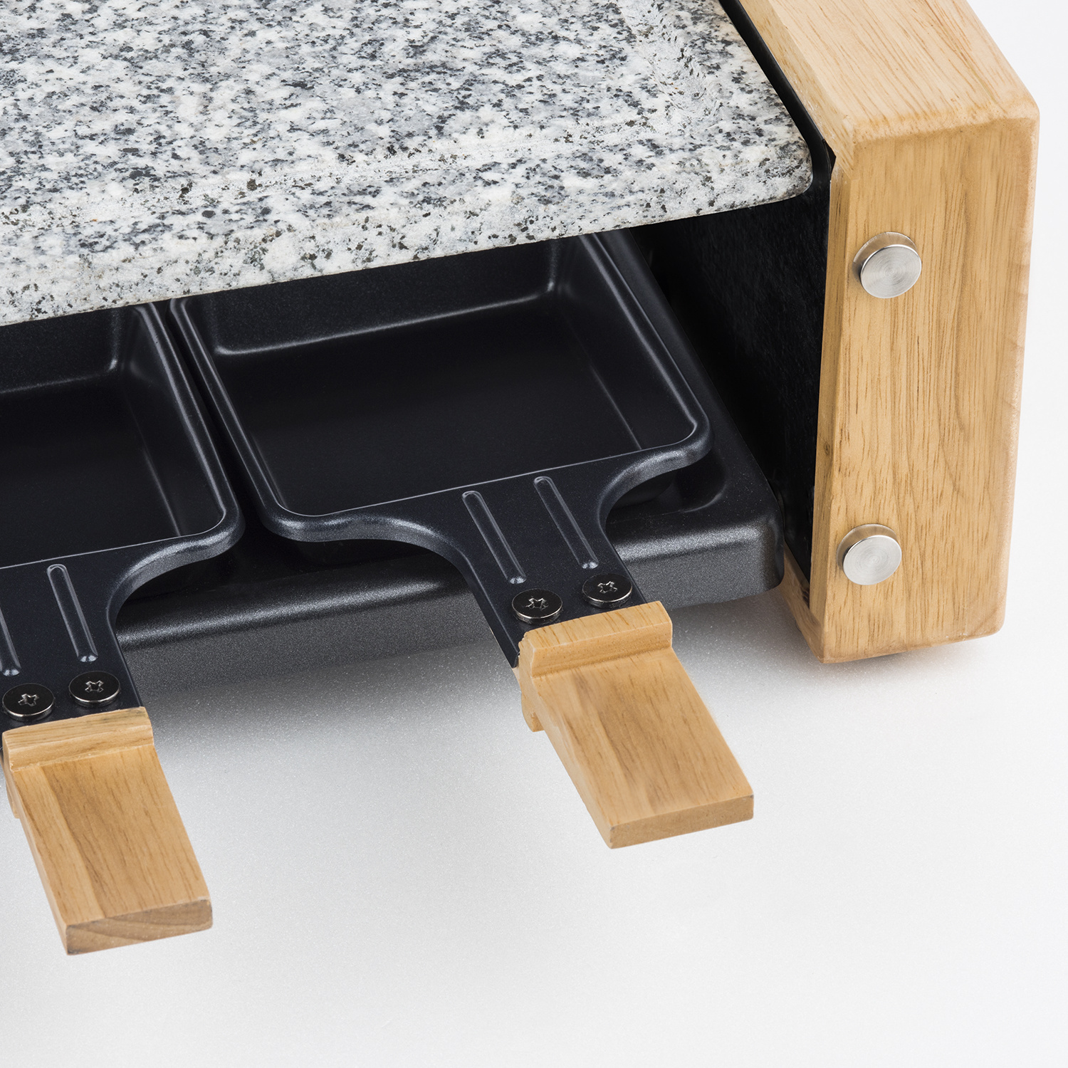 Una raclette in legno con piastra in granito e due mini padelle nere con manici in legno.