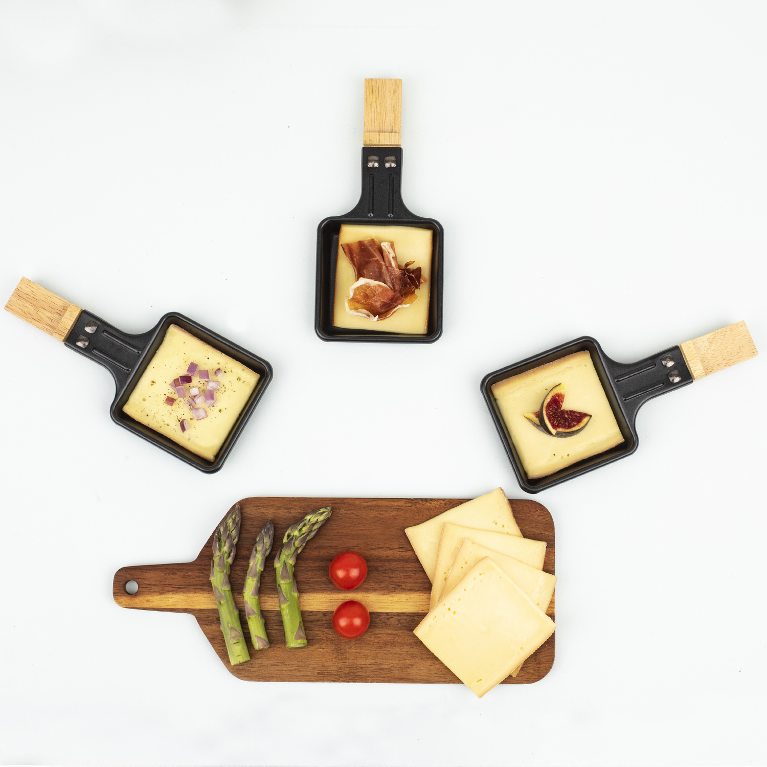 Tre padelle per raclette con formaggio, prosciutto, fichi e cipolle, e un asse di legno con asparagi, pomodori e formaggio.
