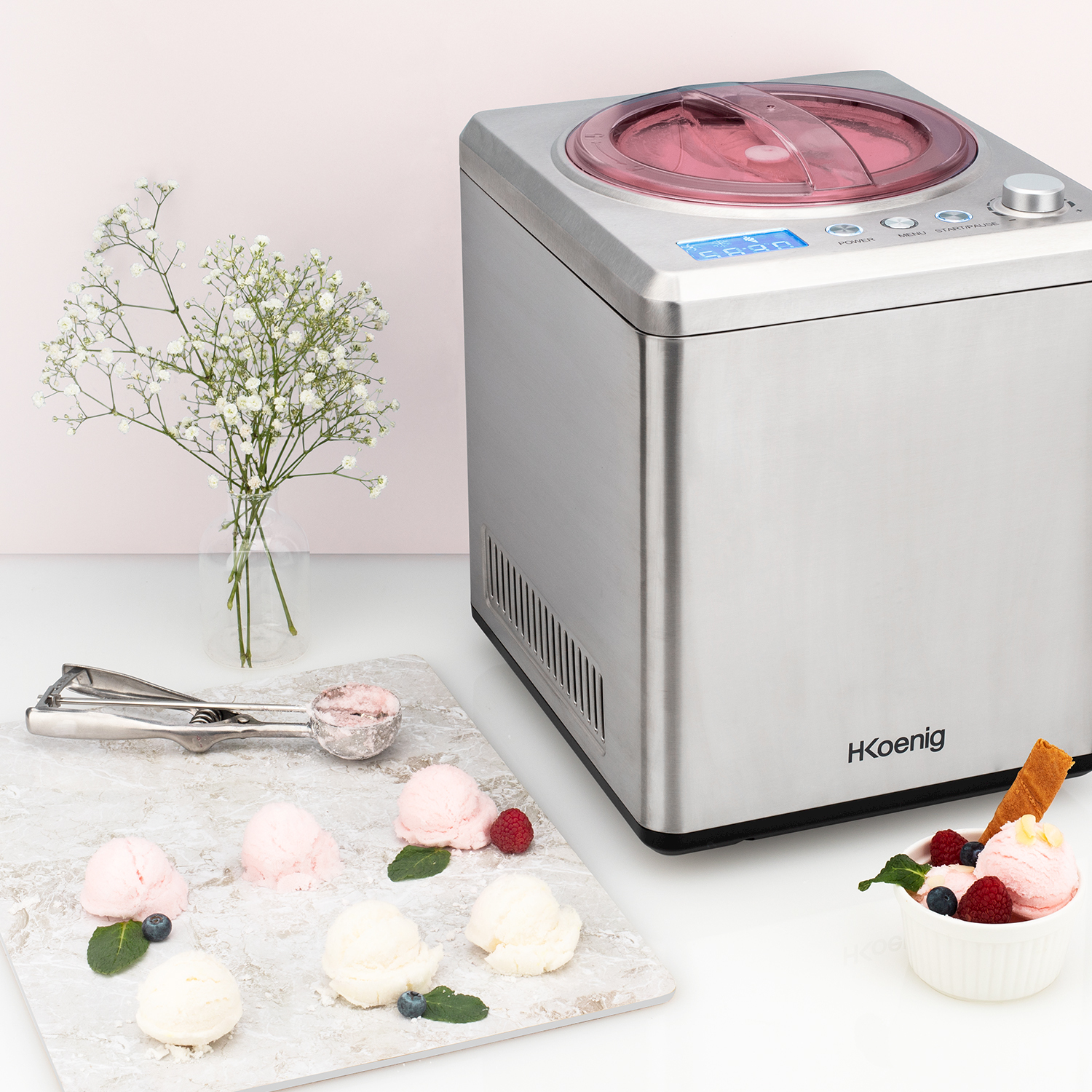 Una gelatiera in acciaio inossidabile si trova accanto a palline di gelato, bacche e fiori.