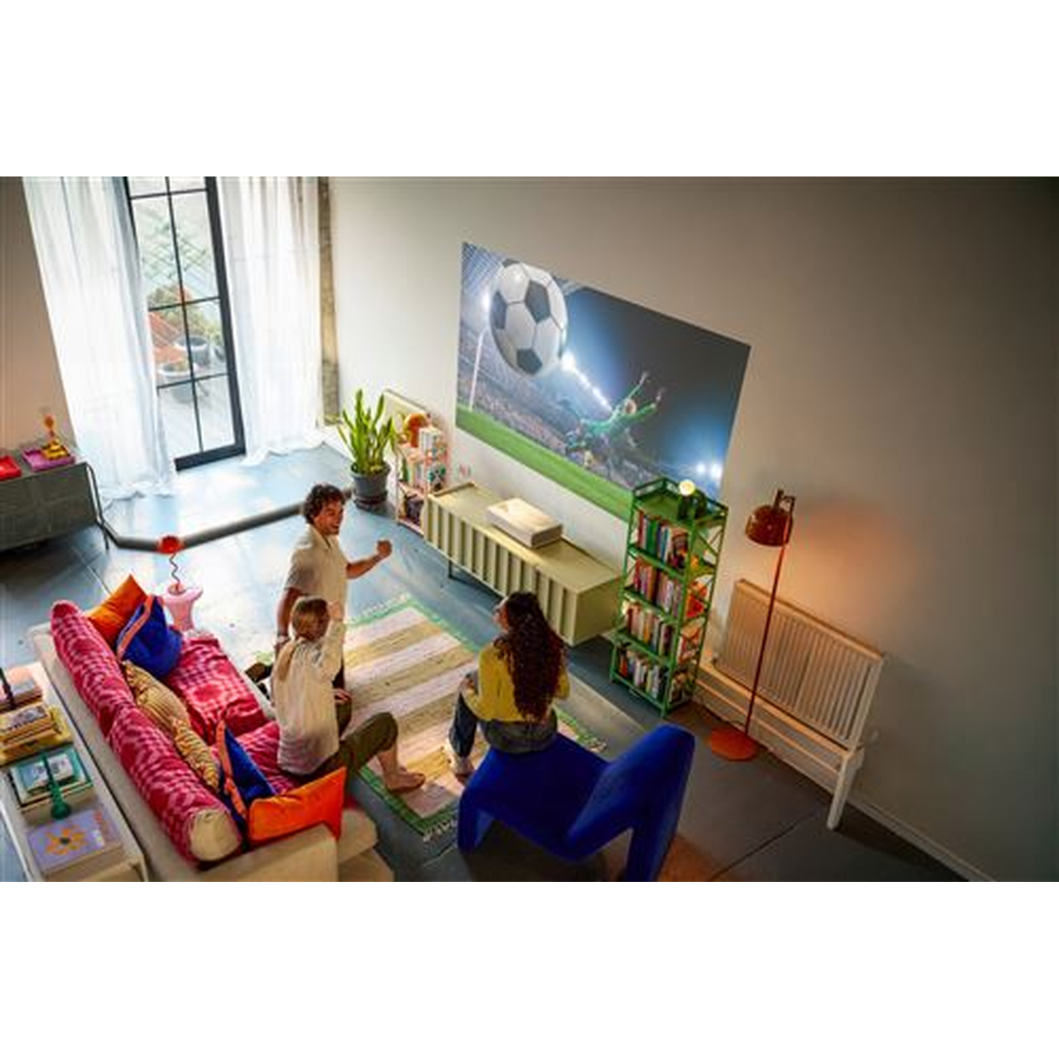 Drie personen kijken naar voetbal op een groot projectiescherm in een woonkamer.
