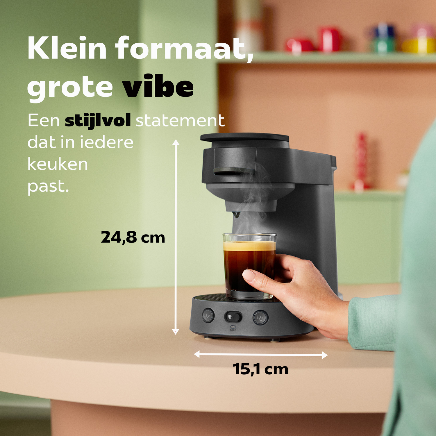 Een hand houdt een glazen kop koffie vast onder een zwarte koffiemachine met afmetingen.