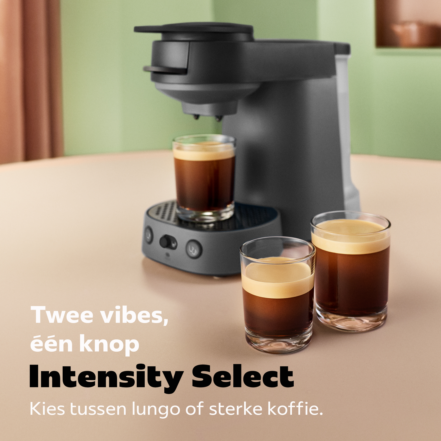 Een grijze koffiezetapparaat op tafel met drie glazen verse koffie, met Nederlandse tekst ervoor.