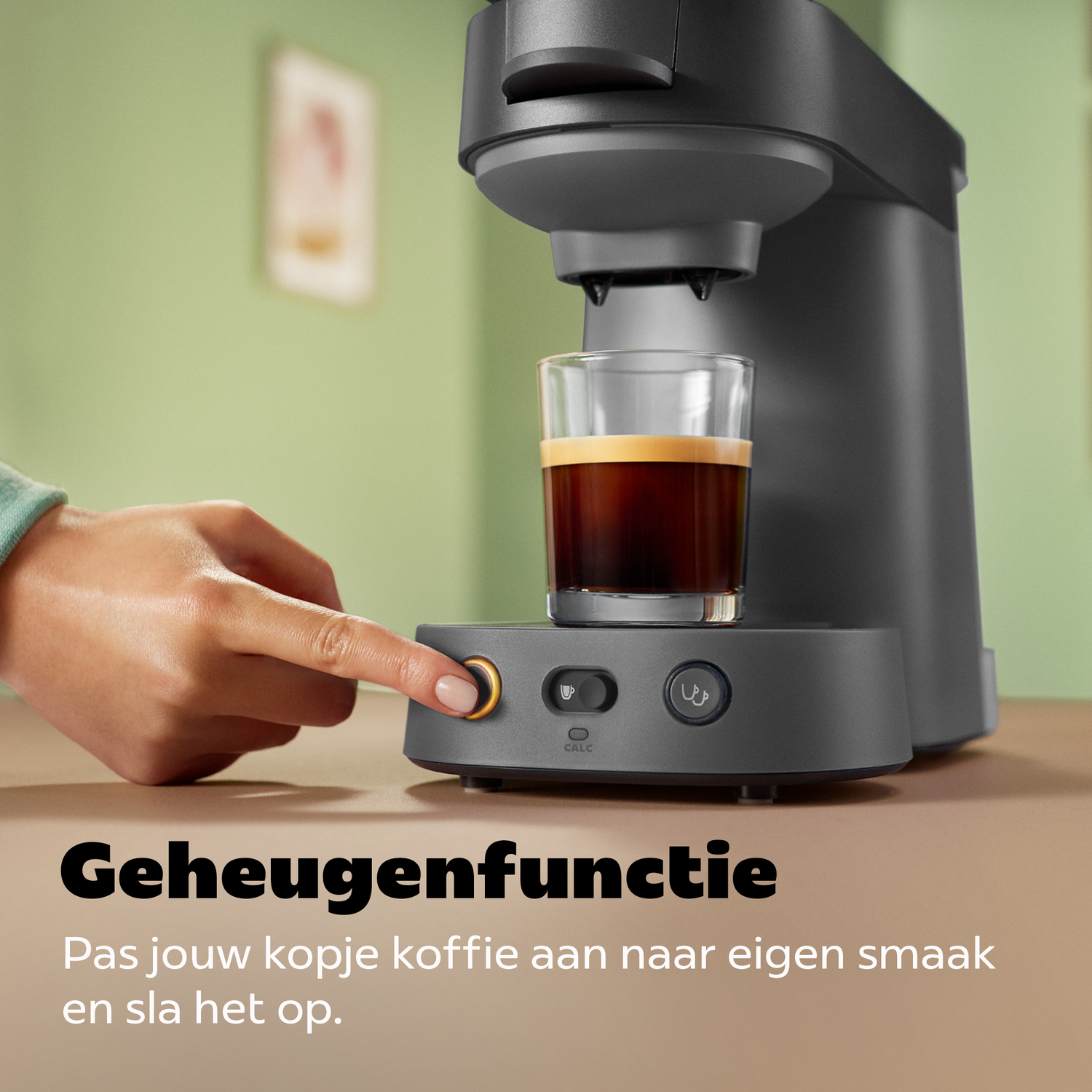 Een hand drukt op de startknop van een grijs koffiezetapparaat met een glazen kop koffie.