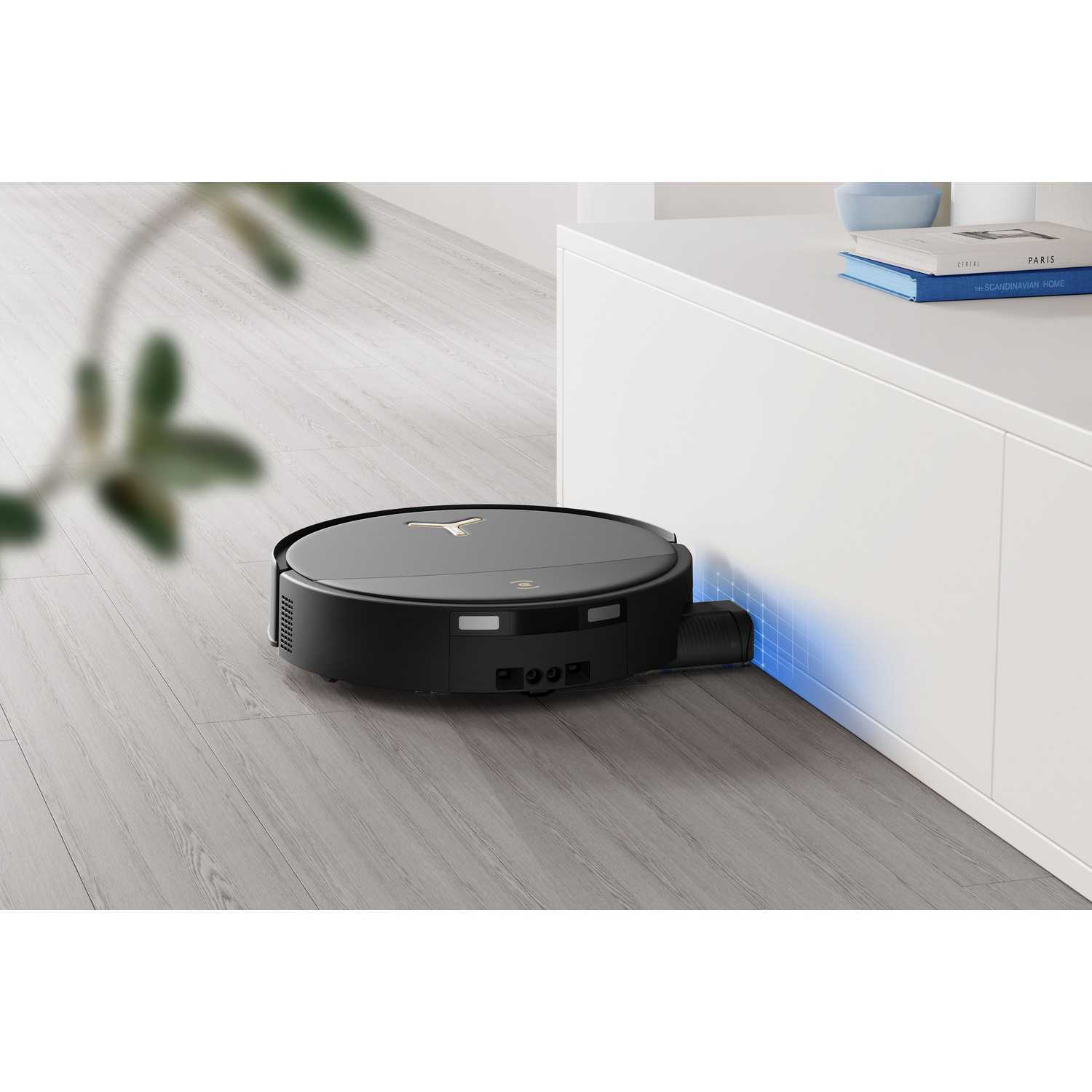 Un robot aspirapolvere nero pulisce il pavimento in una stanza con pavimento in legno e un mobile bianco.