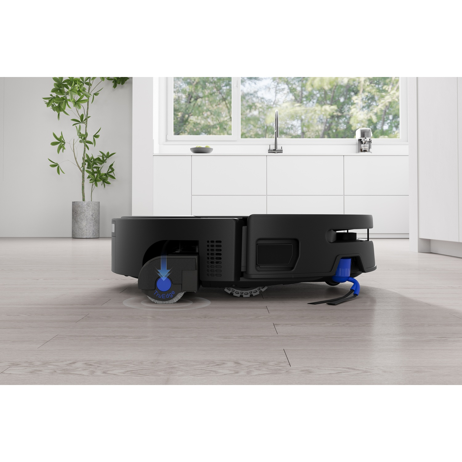 Un robot aspirapolvere nero su un pavimento in legno chiaro in una cucina moderna con mobili bianchi e una pianta.