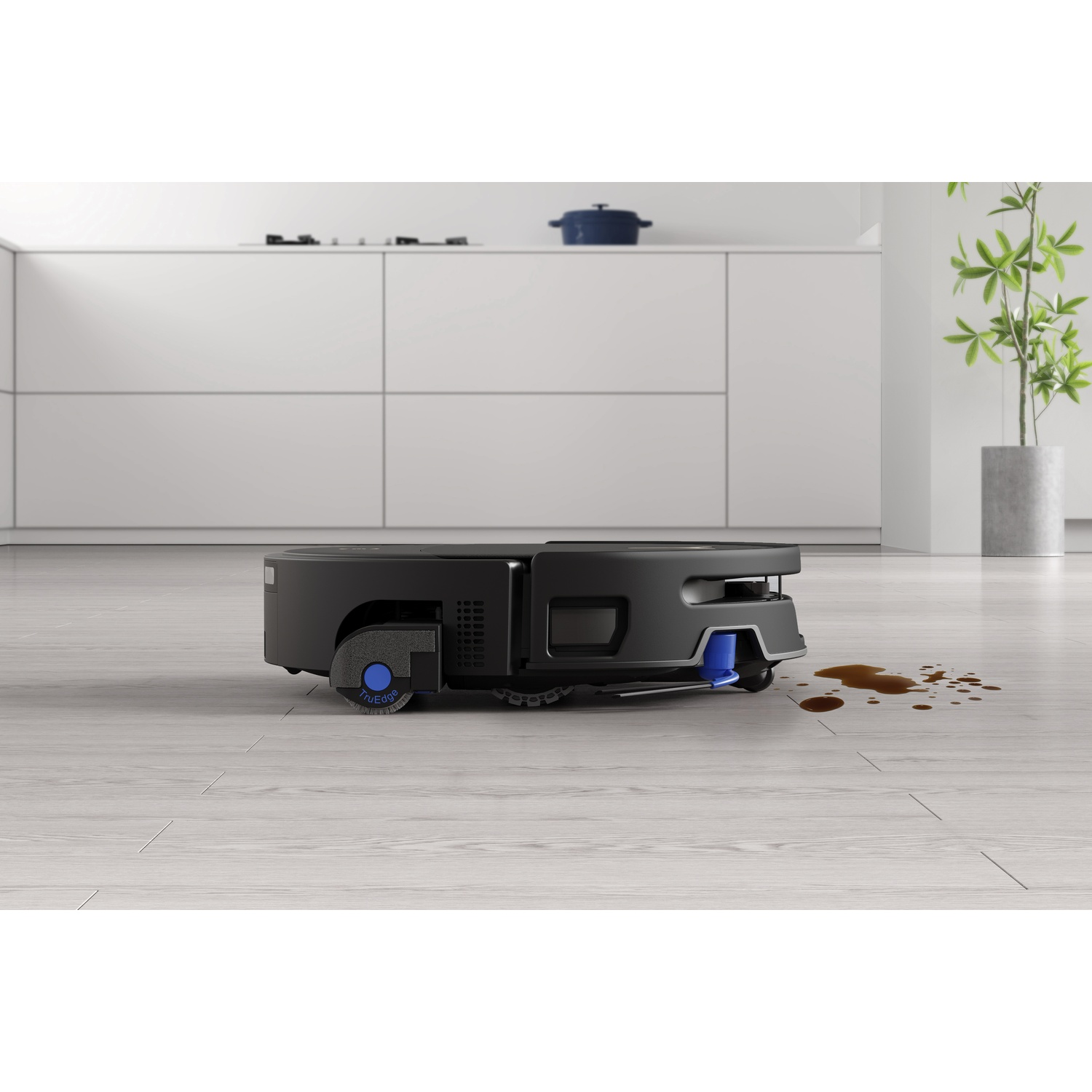 Un robot aspirapolvere nero si avvicina a una macchia di caffè su un pavimento in legno in una cucina moderna.