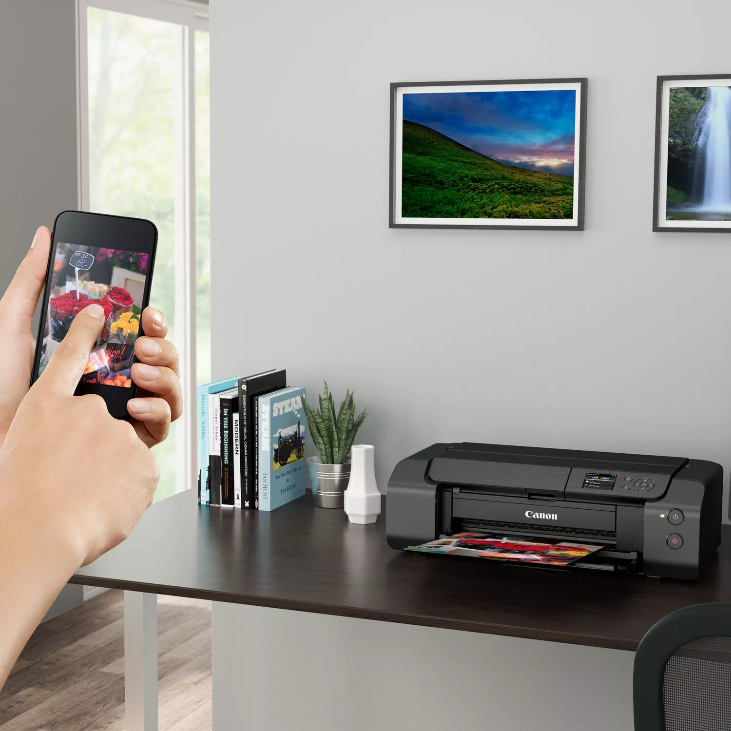 Osoba fotografuje kwiaty smartfonem obok książek i drukarki Canon na biurku.