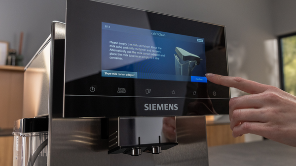 Bir parmak, temizlik uyarısı gösteren bir Siemens kahve makinesinin dokunmatik ekranına dokunuyor.