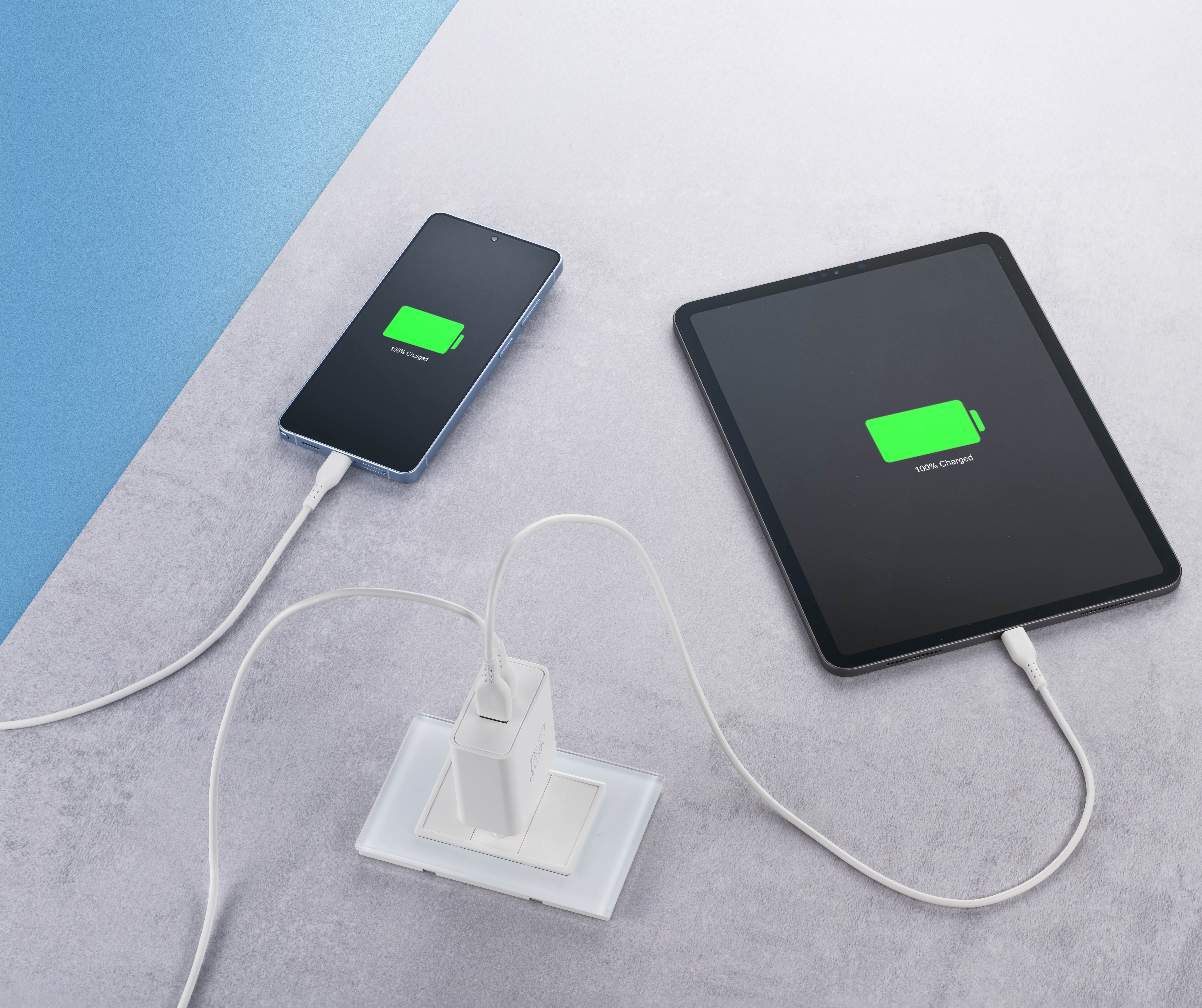 Ein Smartphone und ein Tablet sind an ein weißes USB-Ladegerät in der Steckdose angeschlossen und zeigen 100% Akku.