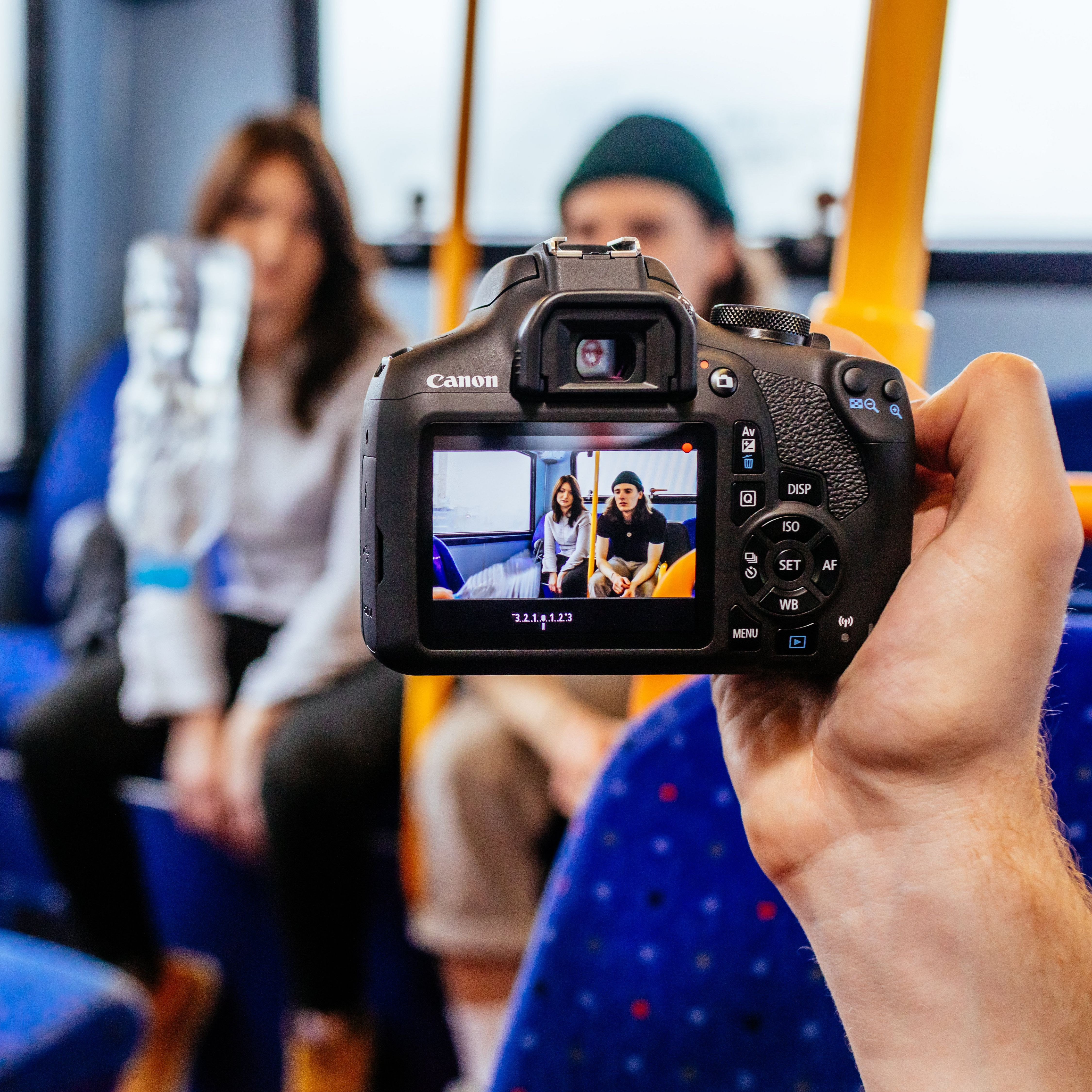 Une main tient un appareil photo Canon affichant deux personnes dans un bus.