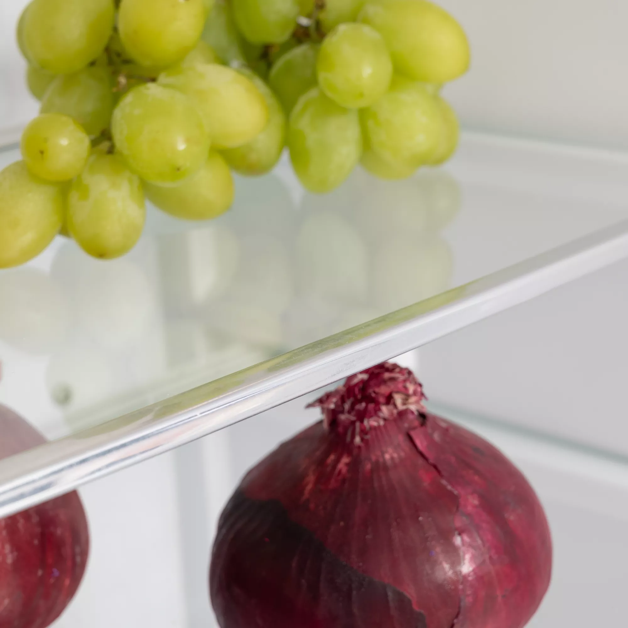 Un racimo de uvas verdes en el estante superior y cebollas rojas en el estante inferior dentro de un refrigerador.