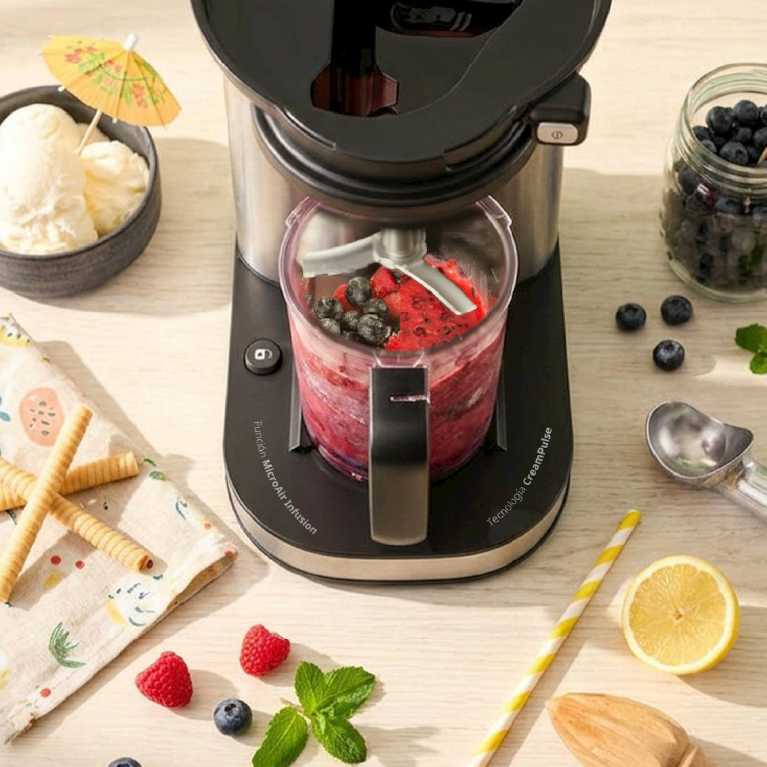 Moderna gelatiera con tazza di miscelazione piena, circondata da frutta, gelato, cialde e accessori su tavolo in legno.