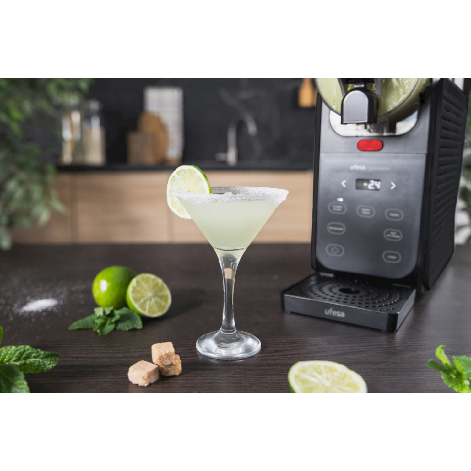 Un bicchiere da cocktail con fetta di lime accanto a una gelatiera nera su un tavolo con lime e menta.