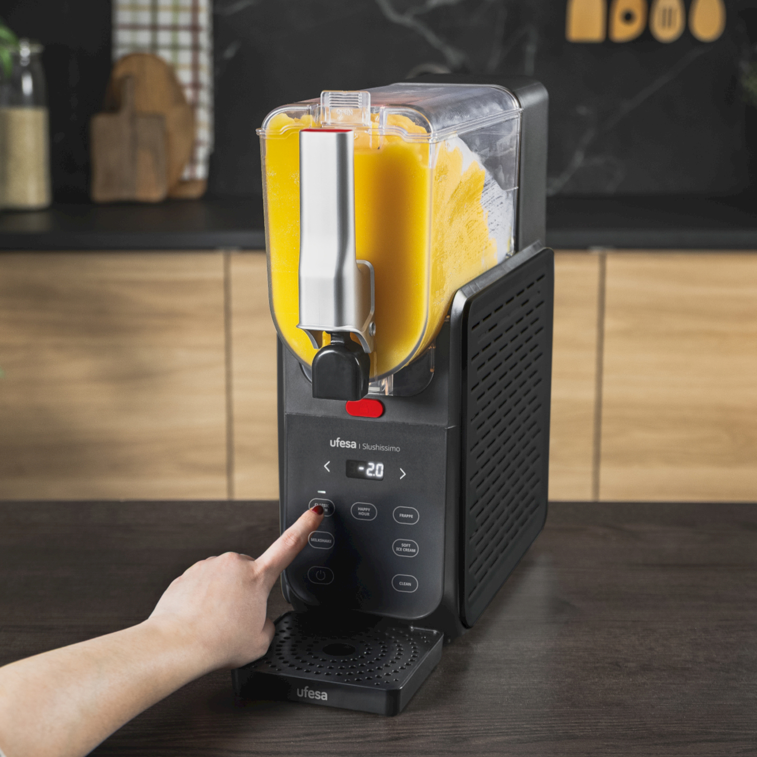 Una mano aziona il display digitale di una macchina per granite nera con contenuto giallo su un tavolo di legno.