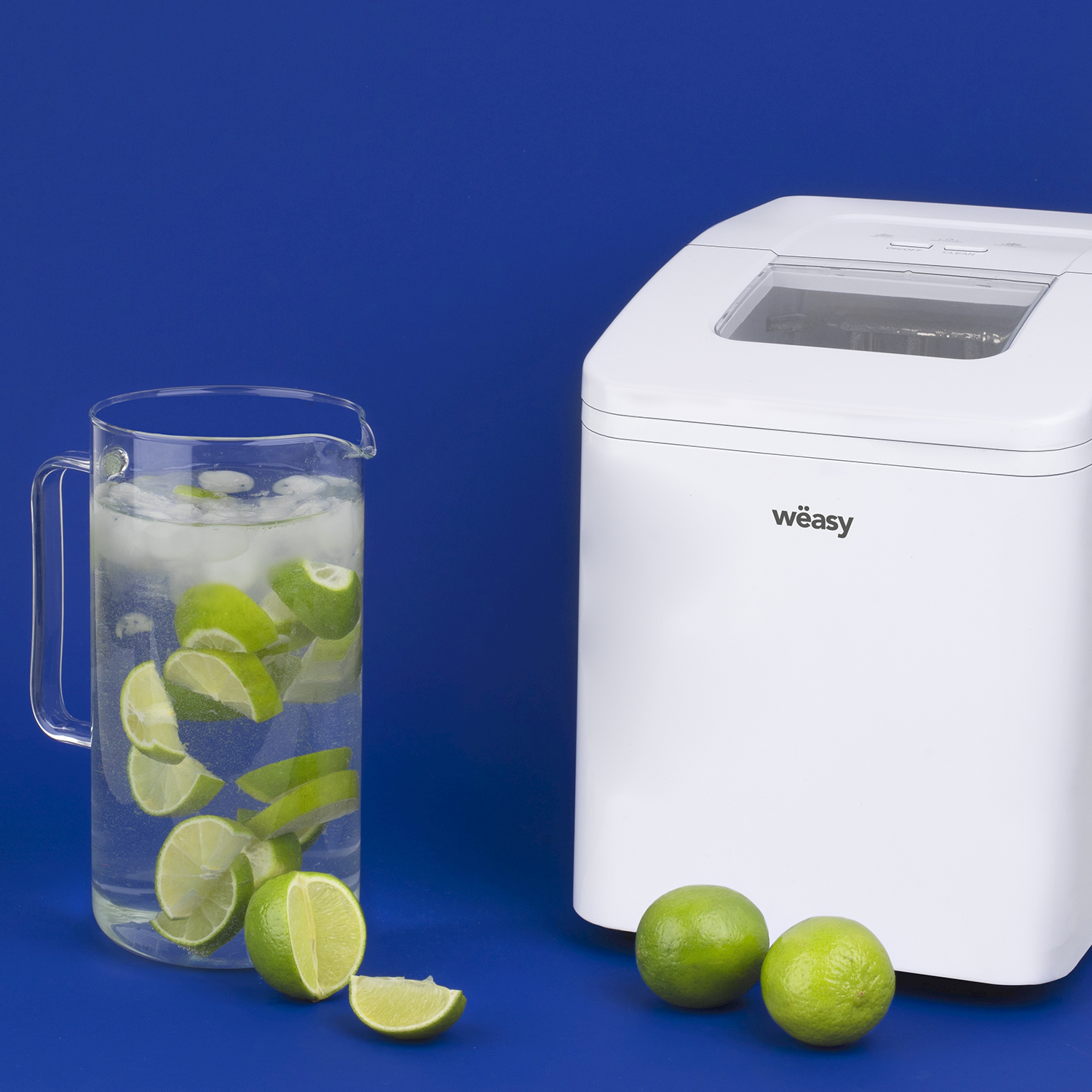 Una macchina per ghiaccio bianca accanto a una brocca di vetro con acqua, ghiaccio e lime su sfondo blu.