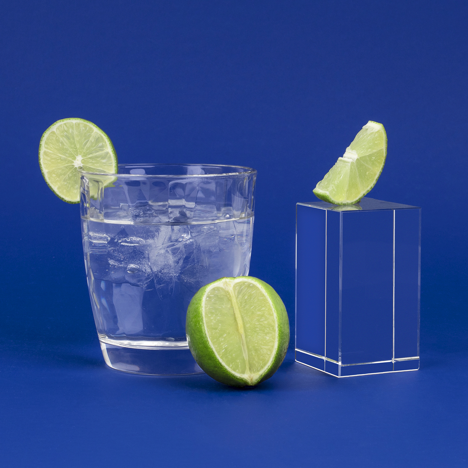 Un bicchiere di acqua ghiacciata con fetta, metà e spicchio di lime su blocco di vetro su sfondo blu.