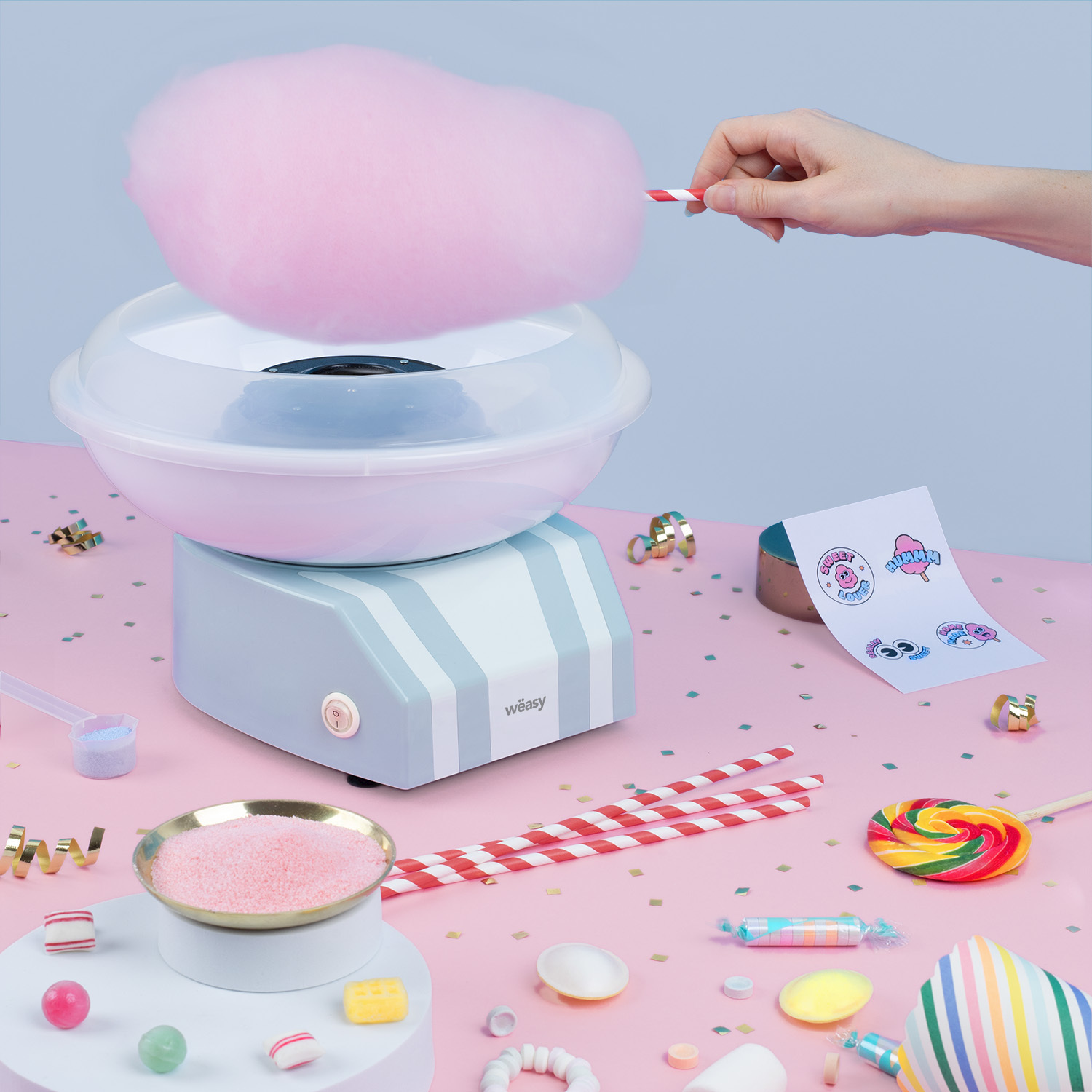 Una mano tiene zucchero filato rosa sopra una macchina azzurra su un tavolo rosa con dolci.