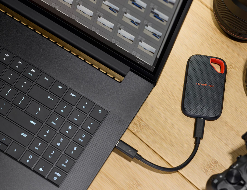 Zewnętrzny dysk SanDisk jest podłączony kablem do czarnej klawiatury laptopa na drewnianej powierzchni.